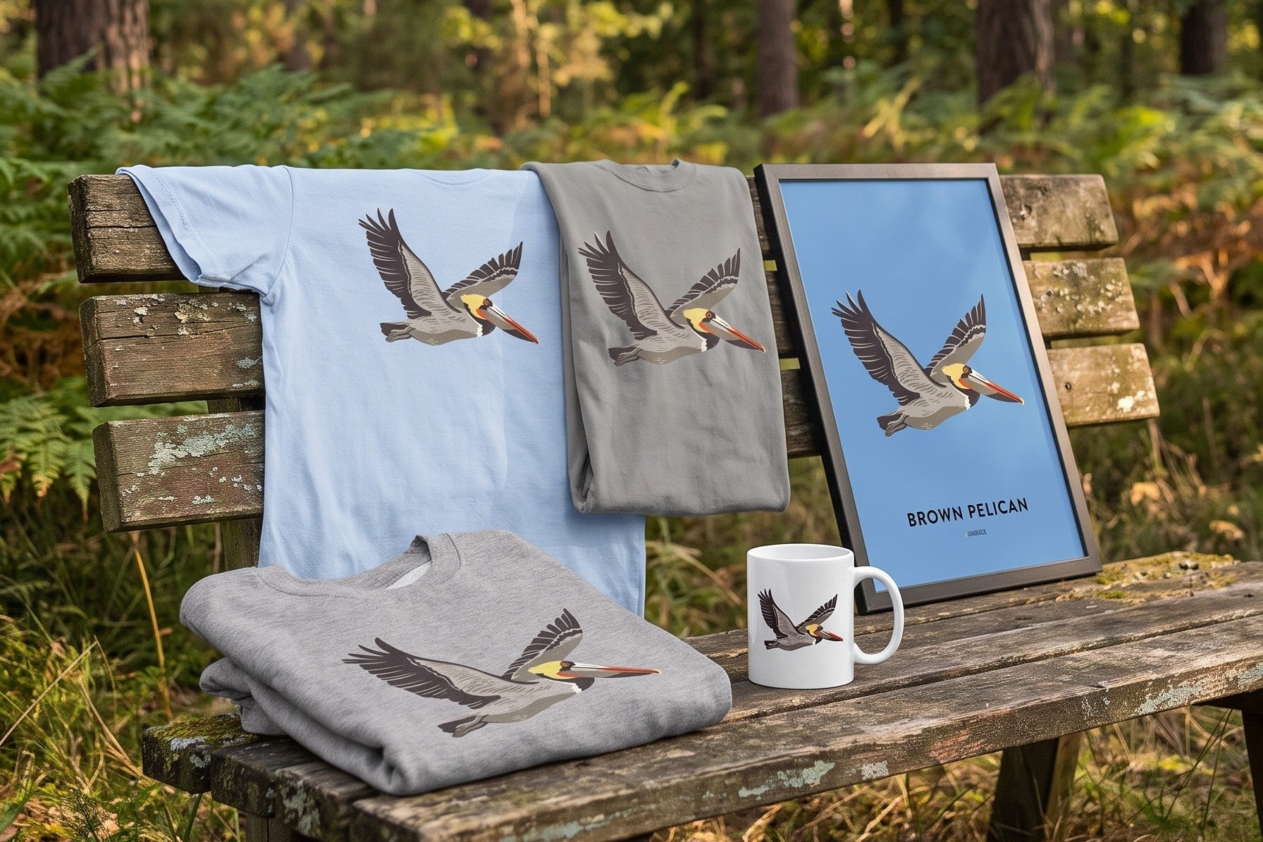Brown Pelican t-shirts, sweatshirt, mug and framed print arranged on a wooden bench in a forest